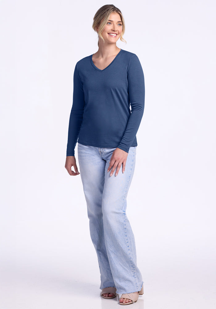 A smiling woman with light hair pulled back poses against a white background, wearing the Woolx Millie Long Sleeve in Starry Night, light blue jeans, and beige open-toe shoes, with one hand resting on her thigh.