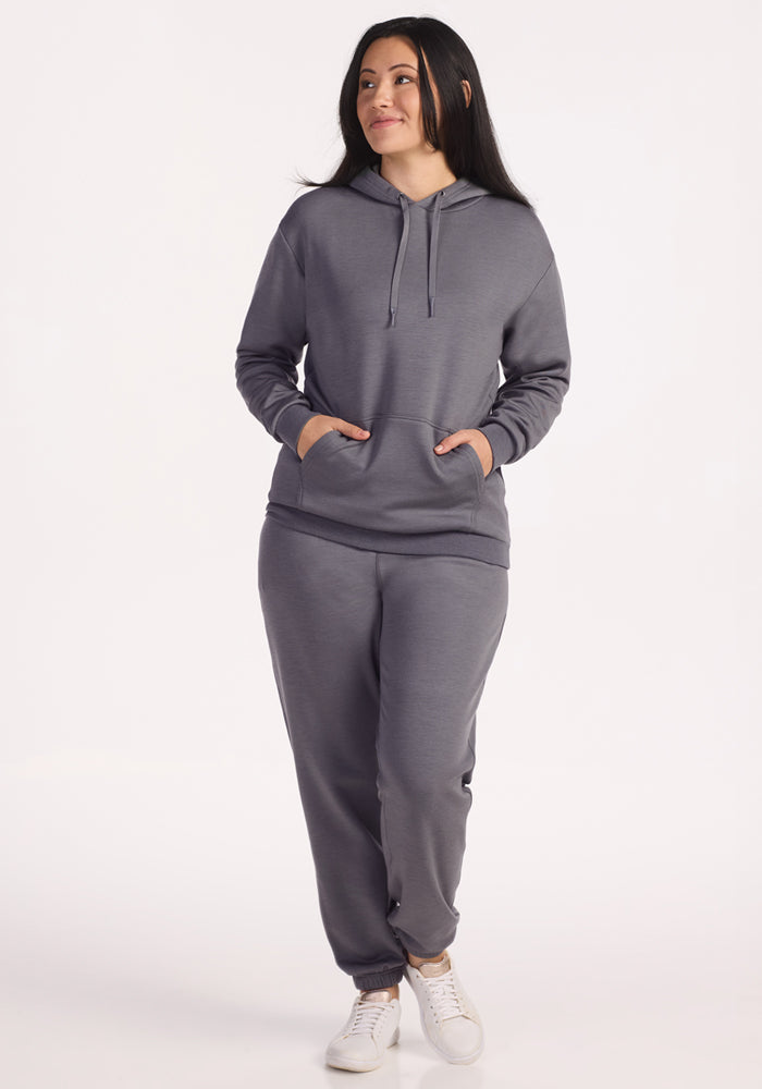 A woman stands smiling with her hands in the pocket of a Woolx Avery Hoodie in Arctic Dusk, paired with matching gray Merino sweatpants and white sneakers, against a plain white background, showcasing stylish year-round comfort.