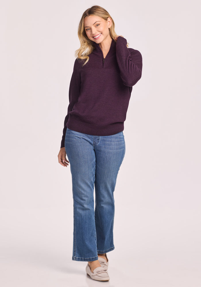 A smiling woman with blonde hair wears the Woolx Collins Half Zip Sweater in Deep Plum, blue jeans, and beige loafers, standing with one hand touching her hair against a plain light background.