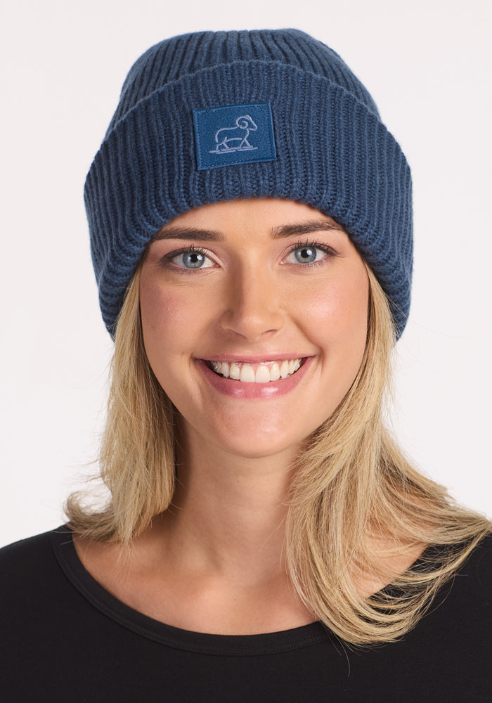 A smiling blonde person wears the Woolx Ria Chunky Beanie - Starry Night, a cozy blue knit hat with a dog patch, standing out against a plain white background.