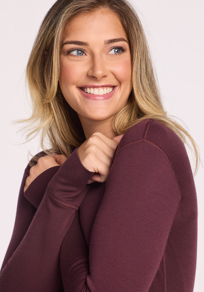 A smiling woman with long, blonde hair wears the Woolx Hannah Long Sleeve Top in Chocolate Fig, holding her hands near her face in a cozy gesture. The moisture-wicking merino wool keeps her comfortable against a light background.