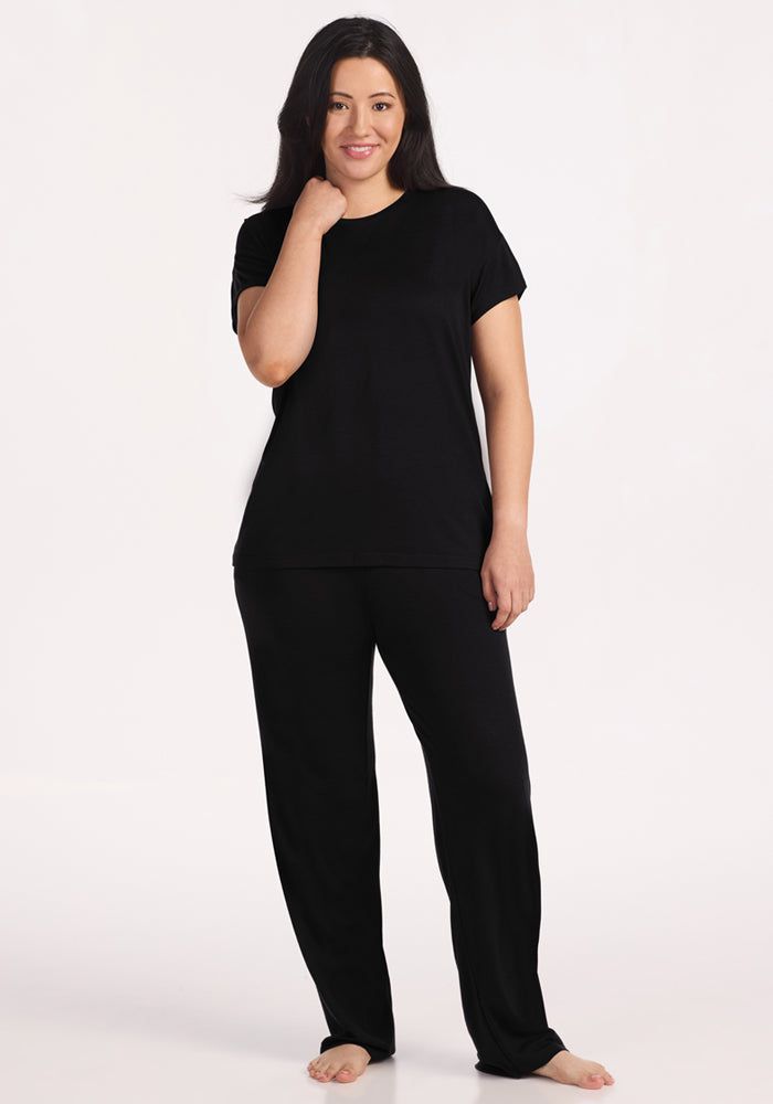A woman with long dark hair stands barefoot on a light background, smiling and touching her neck while wearing the Woolx Billie Lounge Tee in Black with matching loose black pants.