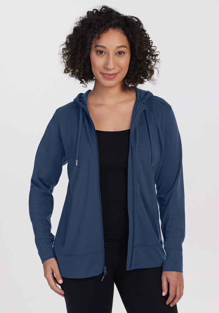 A woman with curly hair, wearing the Woolx Ryann Hoodie in Starry Night blue over a black top, stands facing the camera and smiles slightly against a plain white background. 