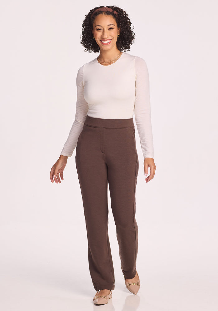 A woman with curly hair smiles against a white background, wearing a long-sleeve white top, Woolx Emerson Straight Leg Pants Petite in Bracken, and beige flats.