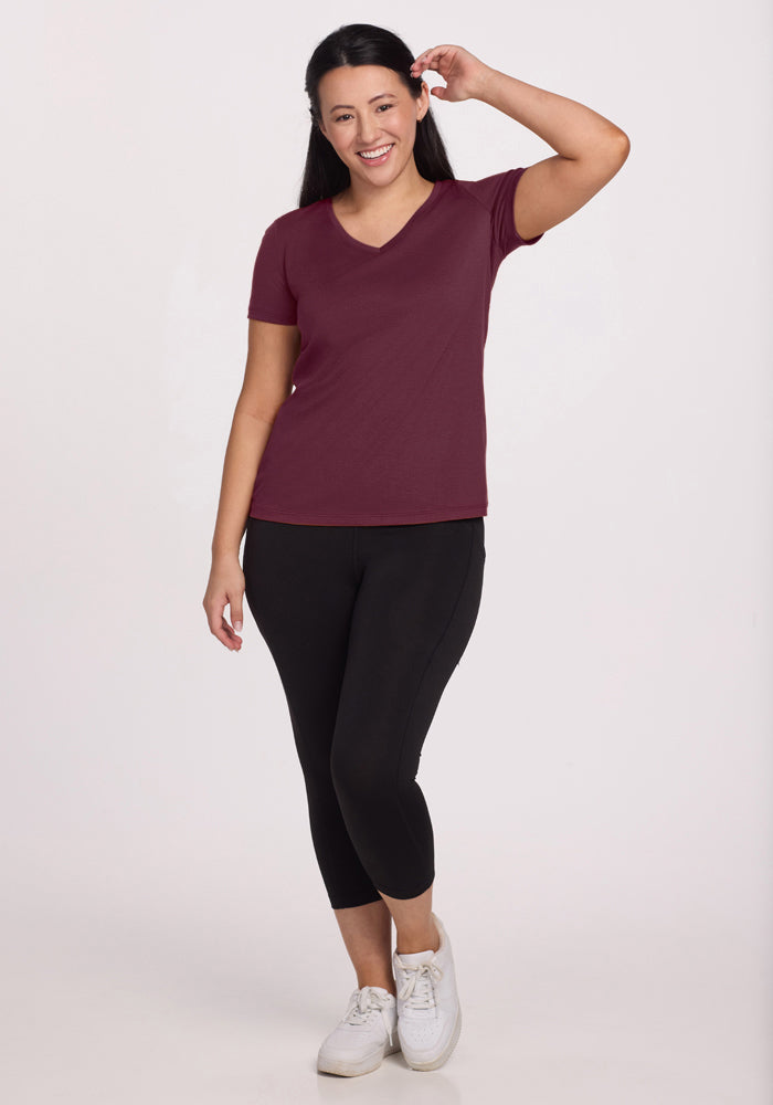 A woman with long dark hair smiles and touches her head while wearing the Woolx Junie V Neck in Grape Harvest, black leggings, and white sneakers against a plain, light background.