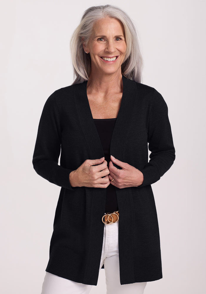 A smiling older woman with long gray hair models the Woolx Macie Sweater in Black over a matching top and white pants, standing against a light background. 