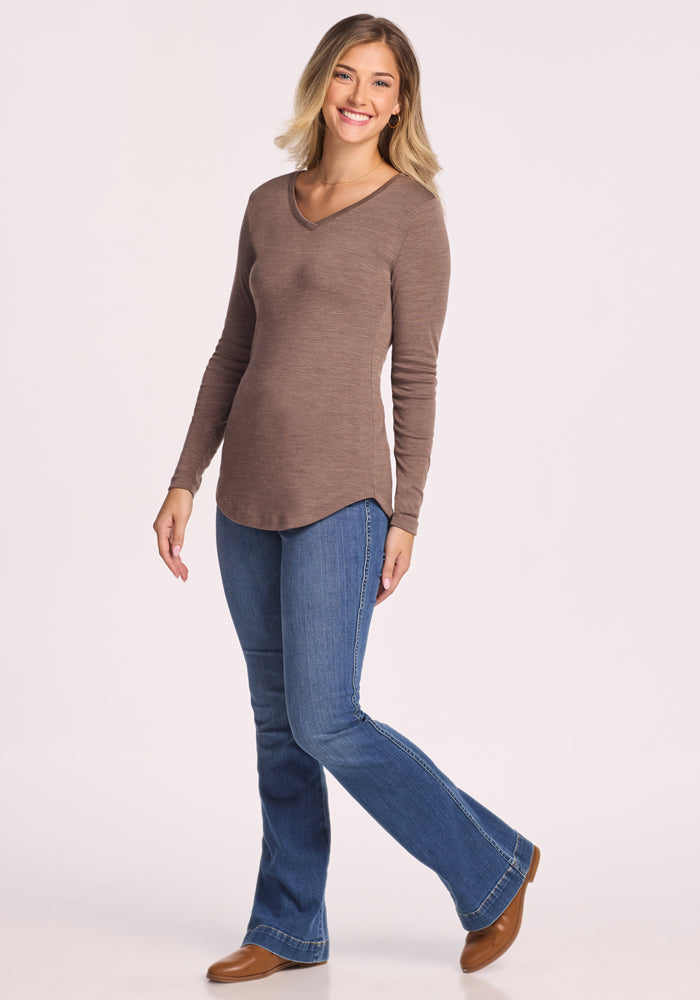 A smiling blonde woman stands against a white background, wearing the Woolx Eva Tunic in Simply Taupe for midweight warmth, paired with blue bootcut jeans and brown shoes.