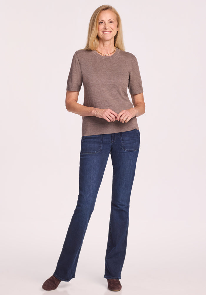 A woman with long blonde hair stands smiling against a plain light background, wearing the Woolx Anna Crew Sweater in Simply Taupe, paired with dark blue jeans and brown shoes, her hands gently clasped in front.