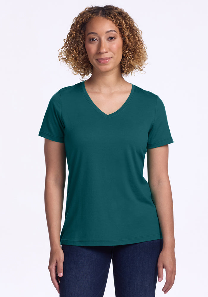 A woman with curly hair wears the Woolx Junie V Neck in Riverbank Green with dark jeans, standing facing forward against a plain white background. 