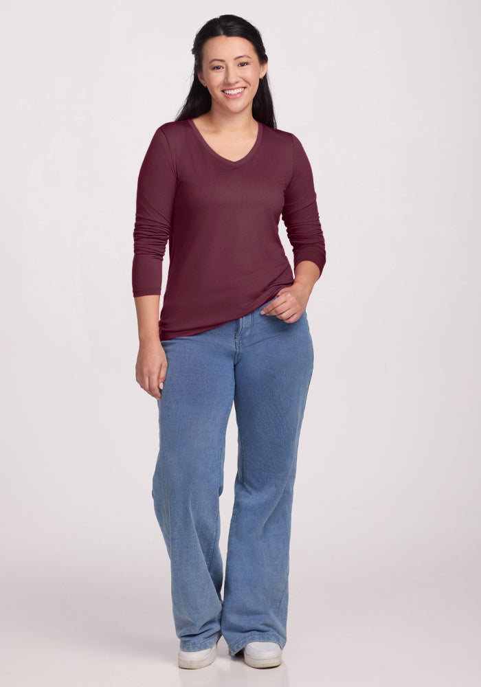 A woman with long, partially tied-back dark hair smiles against a light background, wearing the Woolx Millie Long Sleeve in Grape Harvest. This temperature-regulating, moisture-wicking top pairs with blue jeans and white sneakers for all-day comfort.