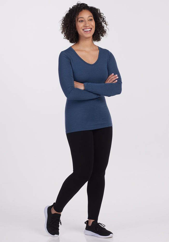 A person with curly hair stands smiling with arms crossed, wearing the Woolx Layla V Neck Top in Starry Night, paired with black leggings and sneakers, against a plain white background.