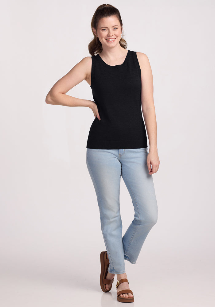 A woman smiles with one hand on her hip, wearing the Woolx Chessy Tank Top in black, paired with light blue jeans and brown sandals against a plain light background—an ideal layering piece for any occasion.