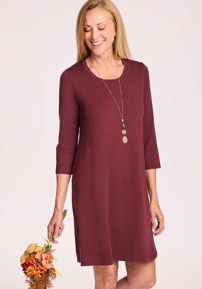 A smiling woman wears the Woolx Rissa A Line Dress in Toasted Garnet with three-quarter sleeves, styled with a long pendant necklace and holding dried flowers—perfect for fall and winter layering against a light background.