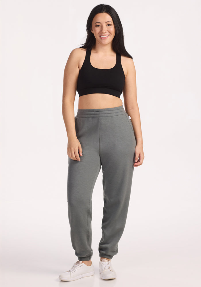 A woman stands smiling against a plain background, wearing a black sports bra, Woolx Parker Sweatpants in Sedona Sage, and white sneakers.