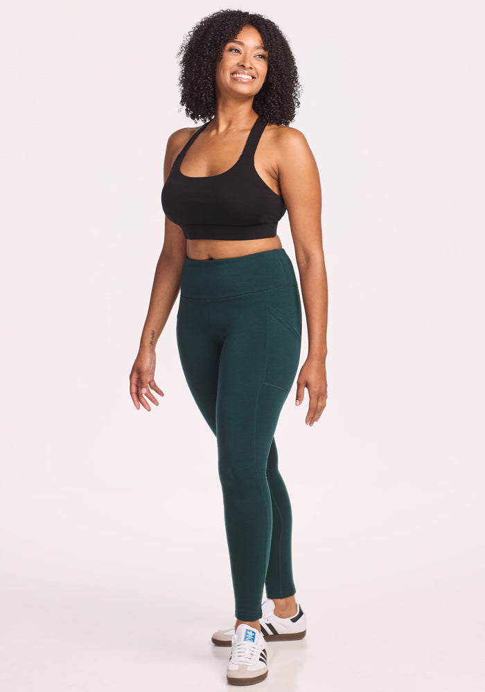 A woman in Woolx’s Piper Pocket Leggings in Deep Timber, a black sports bra, and white sneakers stands smiling and gazing to the side against a plain white background.
