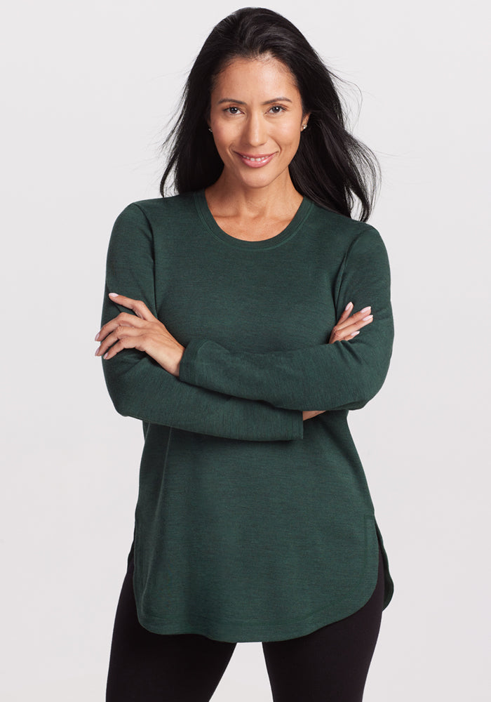 A woman with long dark hair wears the Woolx Blair Tunic in Forest and black pants, standing with arms crossed and smiling against a white background, enjoying lightweight warmth and odor-resistant comfort. 
