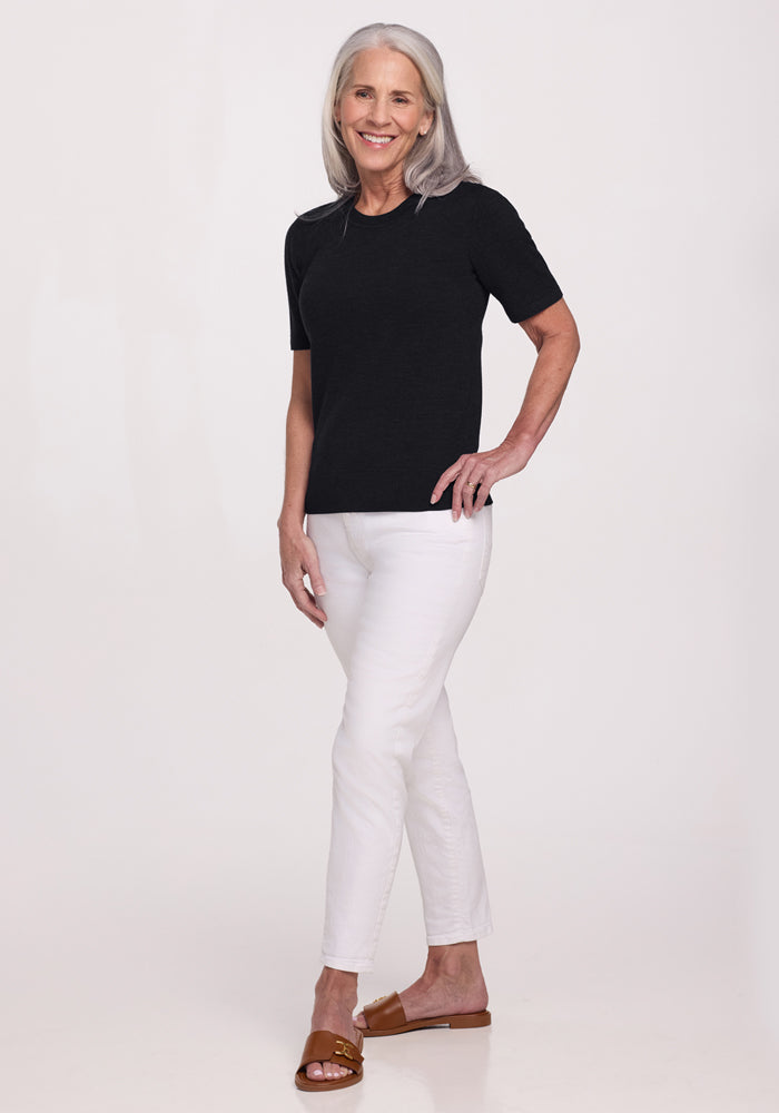 A smiling older woman with long gray hair wears the Woolx Anna Crew Sweater in black, paired with white pants and brown sandals. She stands against a plain light background, one hand on her hip.