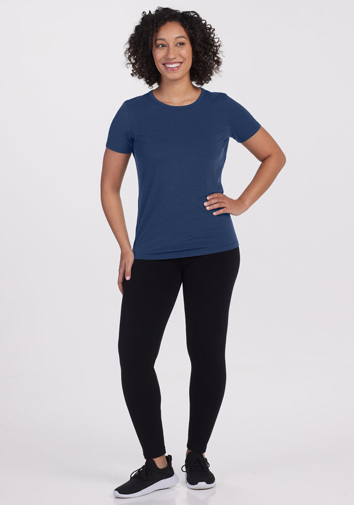 A woman with curly hair smiles against a white background, wearing the Woolx Liza Crew Neck Tee in Starry Night, paired with black leggings and sneakers. One hand rests on her hip, the other at her side.