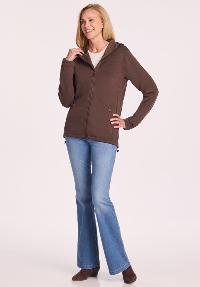 A smiling woman with straight blonde hair models the Woolx Cubby Hooded Sweatshirt in Bracken, paired with light blue jeans and brown boots. She stands against a plain light background, one hand at her chin and the other on her hip.