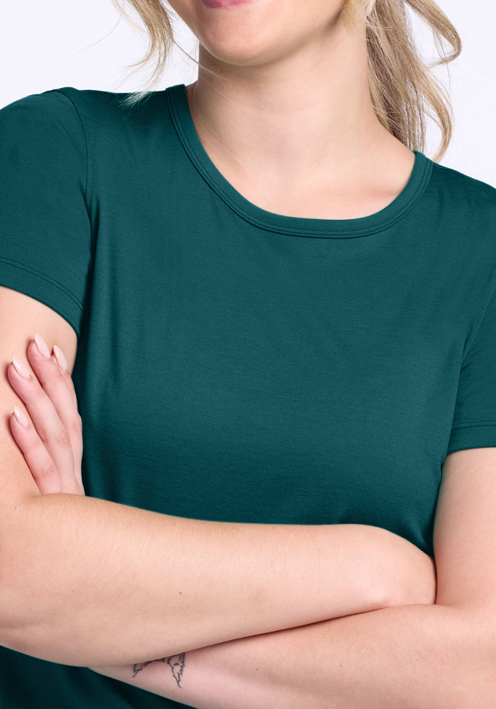 A woman with light skin and blonde hair crosses her arms, wearing the Woolx Liza Crew Neck Tee in Riverbank Green. Only the lower half of her face and upper body are visible.