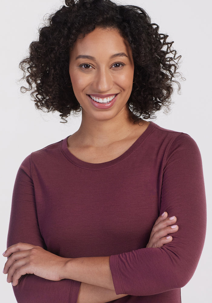 A woman with curly hair smiles confidently at the camera, wearing the Woolx Jenny 3/4 Sleeve Crew Neck in Grape Harvest, arms crossed against a plain light background.