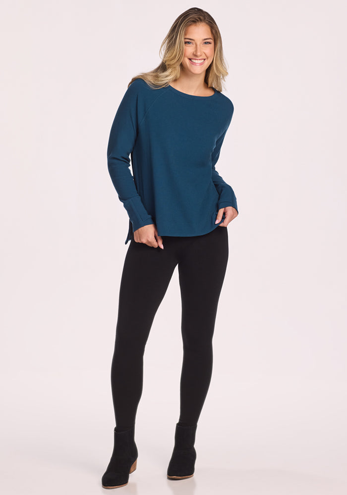 A smiling woman with long blonde hair stands against a plain light background, wearing the Woolx Ainsley Sweater in Majolica Blue, paired with black leggings and black ankle boots.