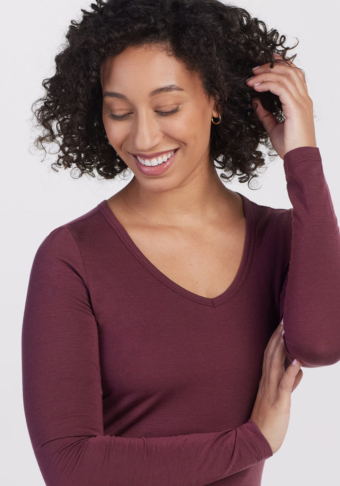 A woman with curly hair smiles, looking down and touching her hair while wearing the Woolx Layla V Neck Top in Grape Harvest against a plain light background.