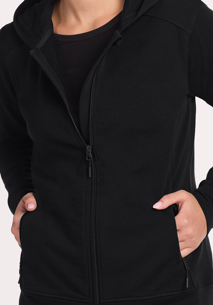 A person wearing the Woolx Darby Zip Up Hoodie - Black stands with hands in the front pockets. The image is cropped from shoulders to hips against a plain, light-colored background.