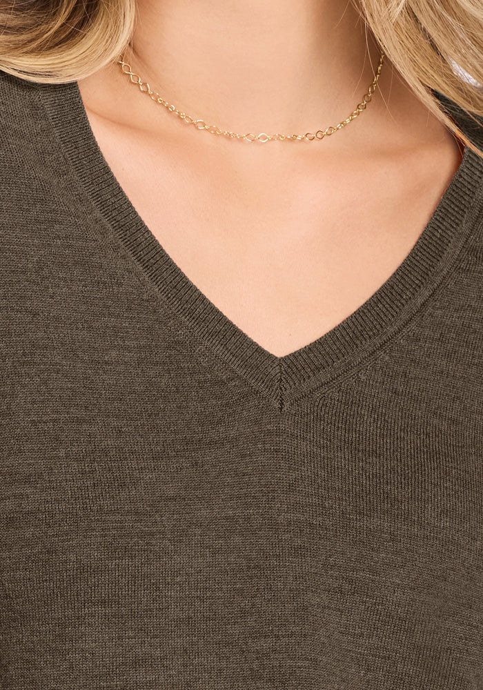 Close-up of a person wearing the Woolx Francesca Sweater in Pine Needle, a dark olive green Merino wool V-neck. A simple gold chain necklace and partially visible hair complete the look.
