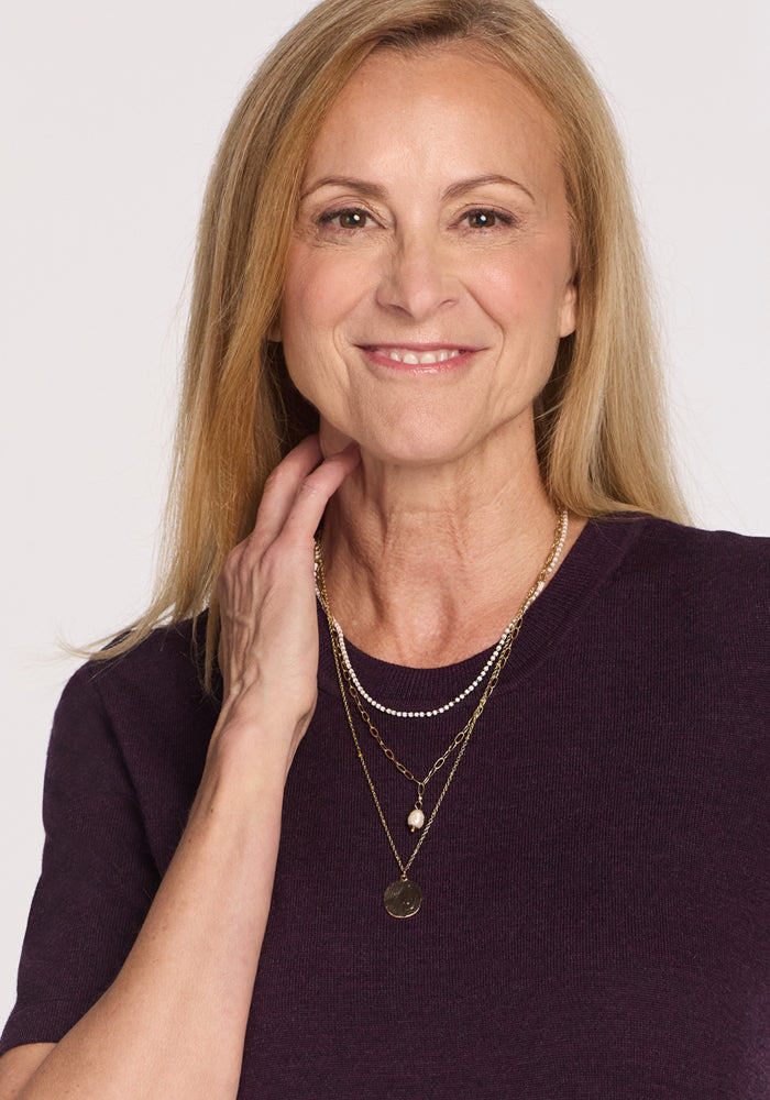 A smiling middle-aged woman with straight blonde hair wears the Woolx Anna Crew Sweater in Deep Plum and layered gold necklaces, posing with one hand to her neck against a plain light background.
