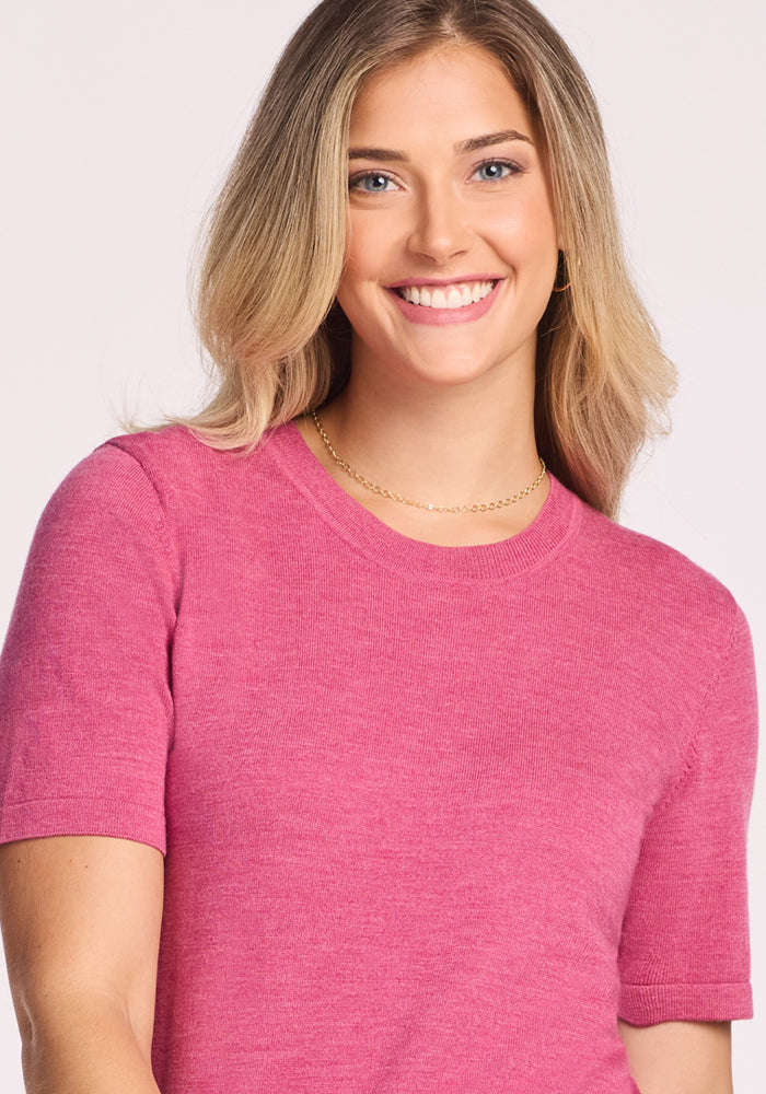 A smiling woman with long, light brown hair models the Woolx Anna Crew Sweater in Wild Salmon and a gold chain necklace, standing against a light background.