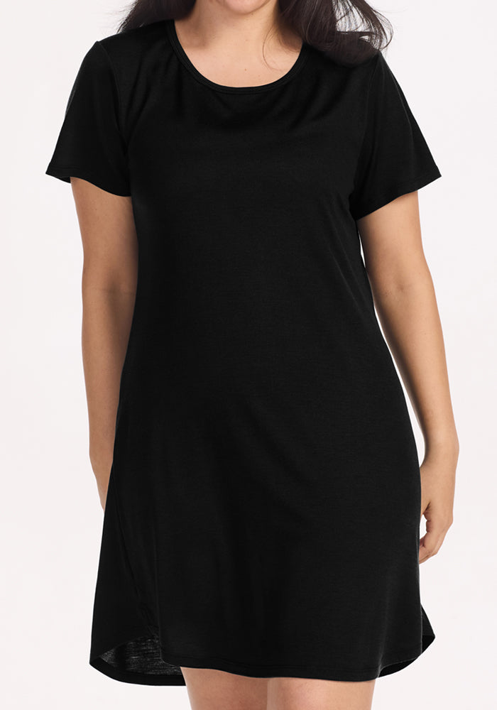 A person models the Woolx Desi Nightgown in black—a short-sleeved, knee-length Merino wool dress with a relaxed fit and round neckline, ideal for temperature regulation as sleepwear or for comfortable travel.