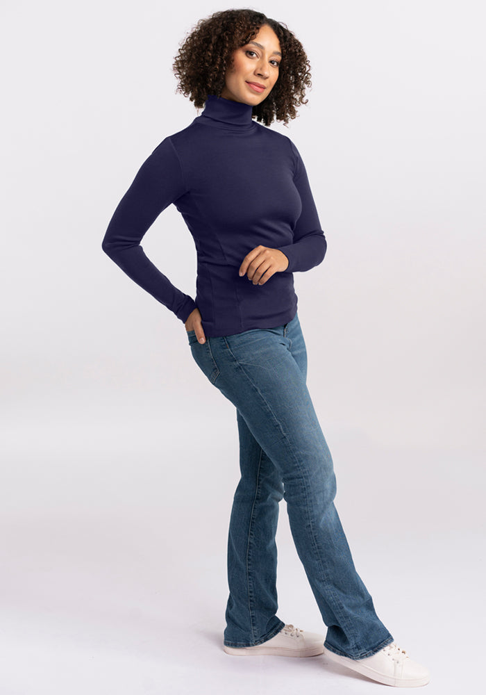 A woman with curly hair in the Woolx Peyton Turtleneck - Midnight Sapphire, blue jeans, and white sneakers stands against a light background, smiling and posing with one hand on her hip.