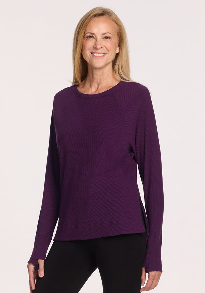 A woman with long blonde hair smiles in a Woolx Ainsley Sweater in Majestic Purple and black pants, standing against a plain light background. 