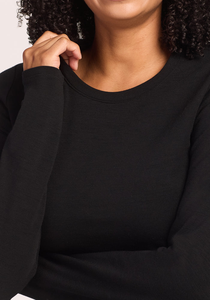 A woman with curly hair, wearing the Woolx Sadie Baselayer Top in Black, touches her collar with one hand; she is smiling and her face is partially visible.