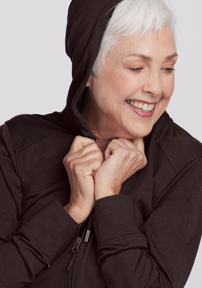 A joyful older woman with short white hair wears the Woolx Ryann Hoodie in French Roast, holding the hood with both hands and looking down in a relaxed pose.