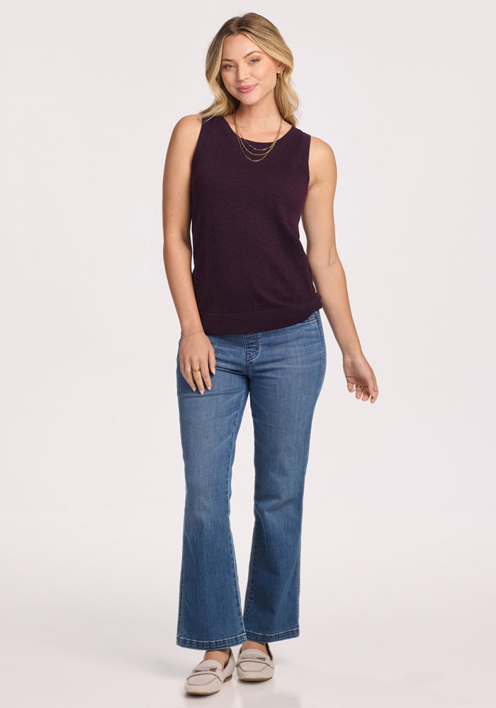 A woman faces forward, smiling softly in the Woolx Chessy Tank Top in Deep Plum, blue jeans, layered gold necklaces, and light-colored loafers. One hand rests on her thigh while the other is by her side.
