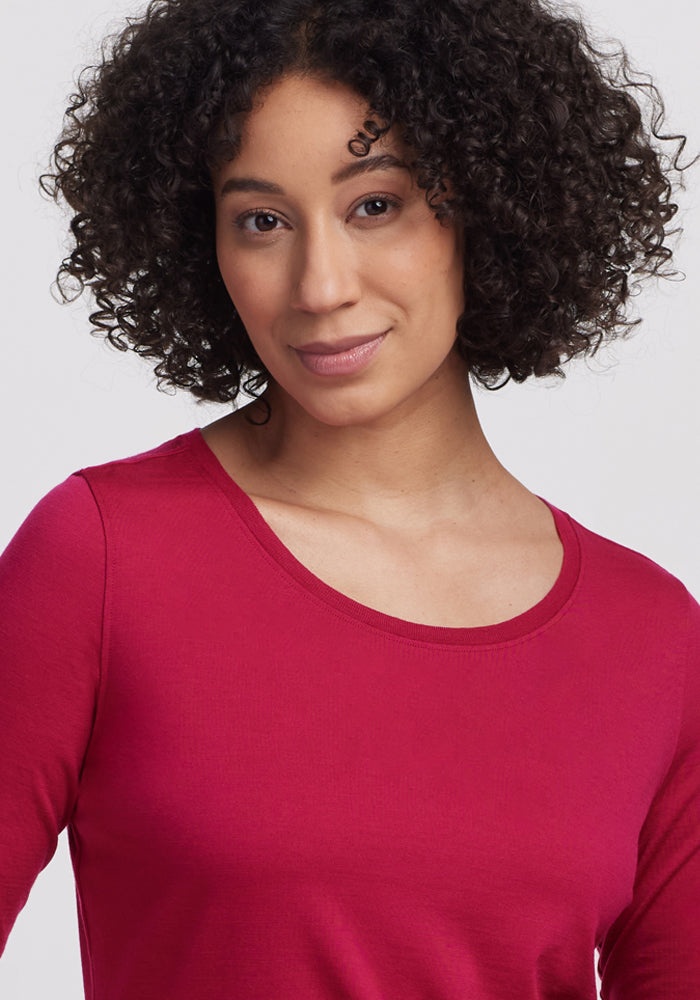 A person with curly dark hair, wearing the Woolx Jenny 3/4 Sleeve Crew Neck in Poppy Spark, poses in front of a plain light background and smiles softly at the camera.