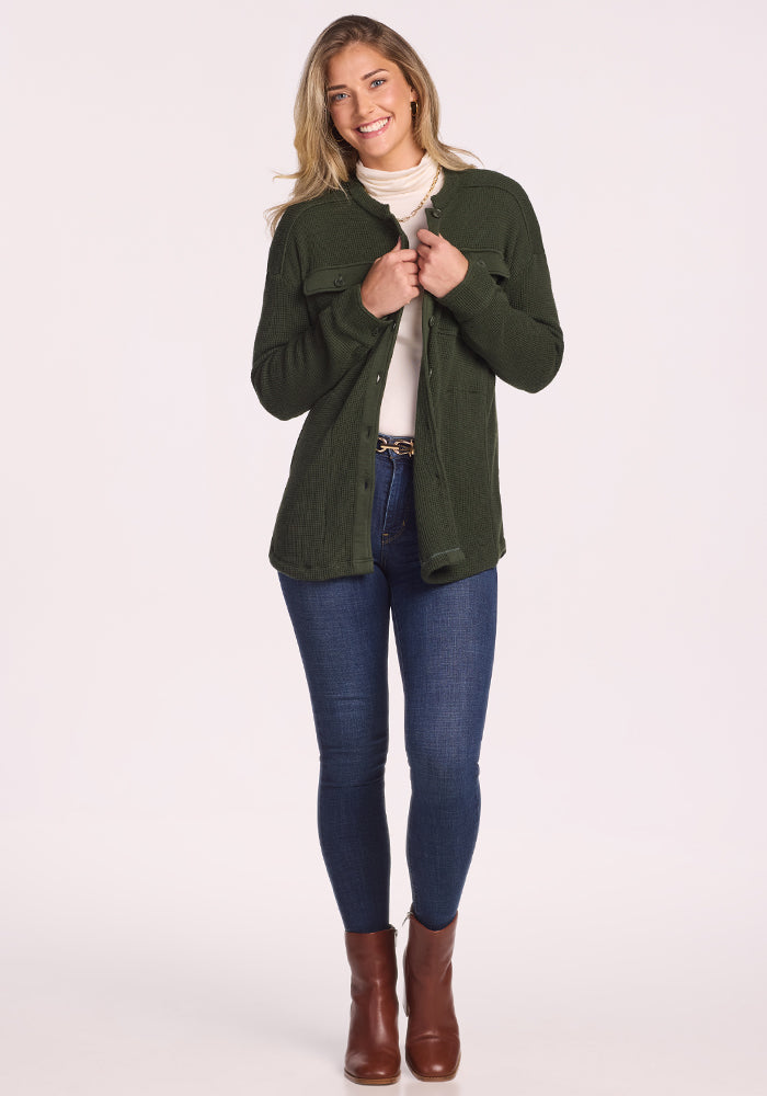 A woman smiles, wearing the Woolx Oaklynn Waffle Shacket in Driftwood Fern over a white turtleneck, blue jeans, and brown ankle boots against a plain light background.