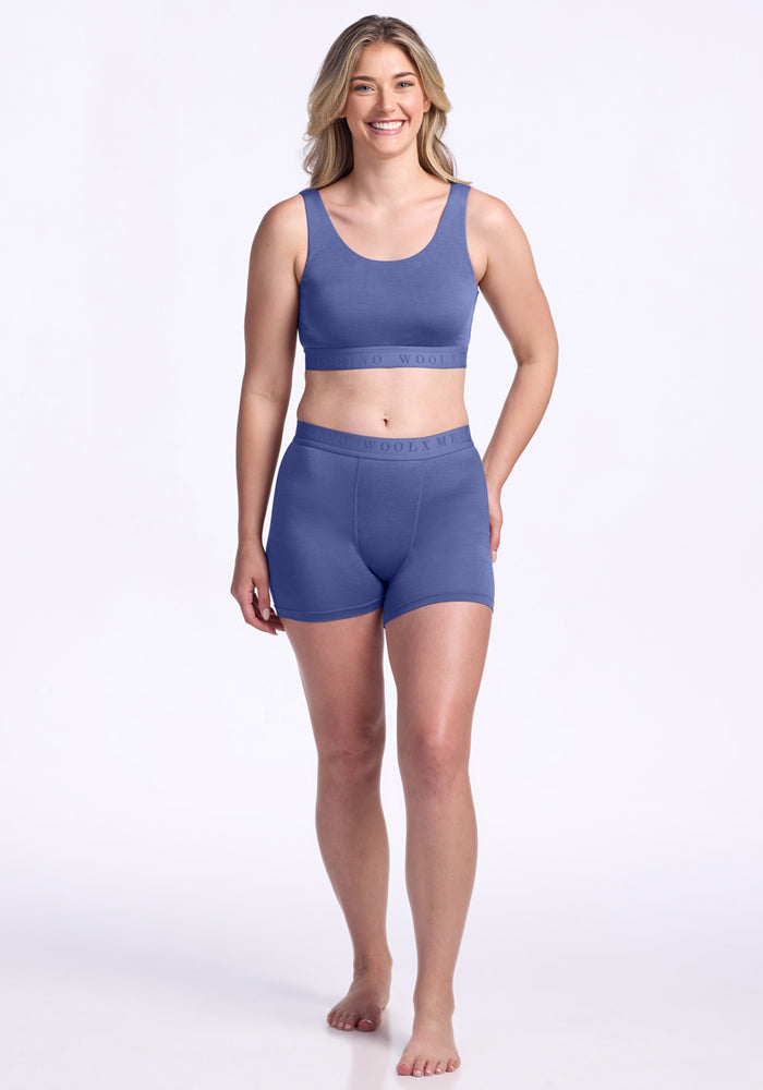 A woman with long blonde hair stands barefoot, smiling, and wearing the Woolx Aubrey Lounge Bra - Cloudy Bay and matching high-waisted boyshorts against a plain light background.