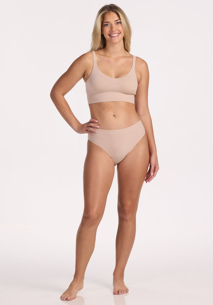 A woman with long blonde hair stands barefoot, smiling, wearing the Woolx Maeve Bikini in Sand—a matching nude-colored bra and high-waisted, moisture-wicking underwear set—against a plain white background.