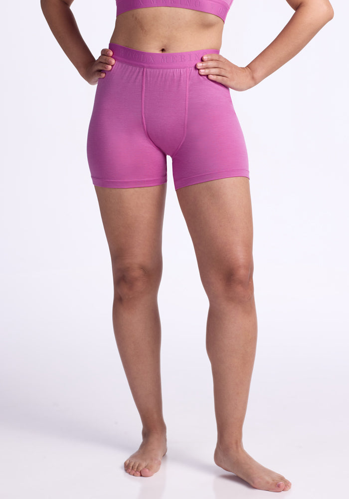 A person, shown from the shoulders down against a white background, models Woolx Elliott Boyshort - Wild Blossom: a quick-drying matching set of pink shorts and crop top. The person is barefoot with arms slightly bent. 