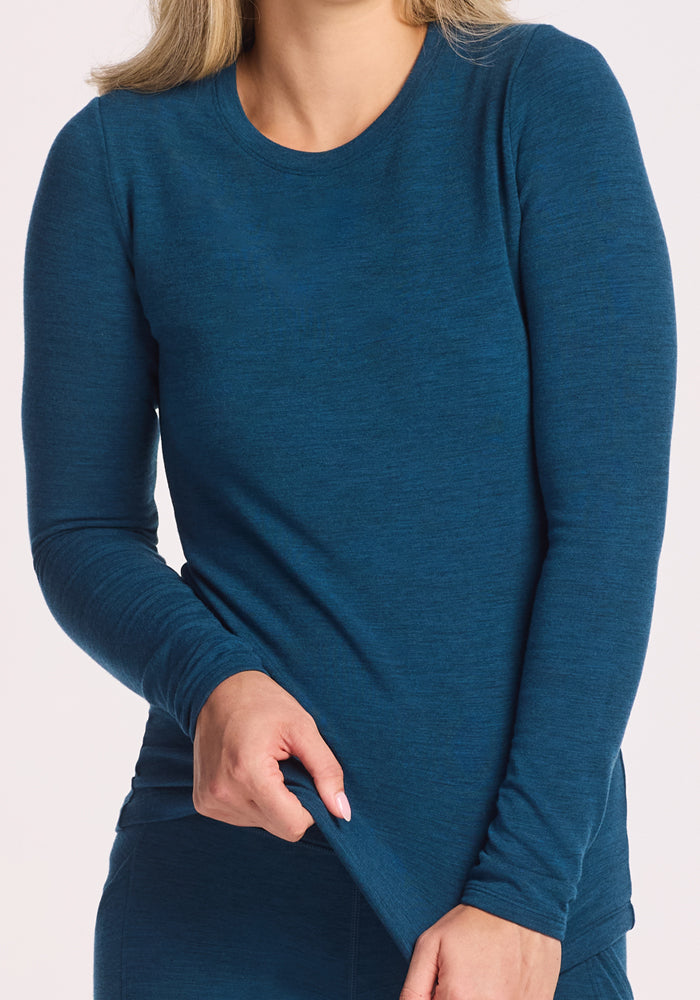 A person in the Woolx Sadie Baselayer Top in Majolica Blue, made from Duralite merino fabric, gently pulls the shirt to the side. The head is partially out of frame against a plain white background.