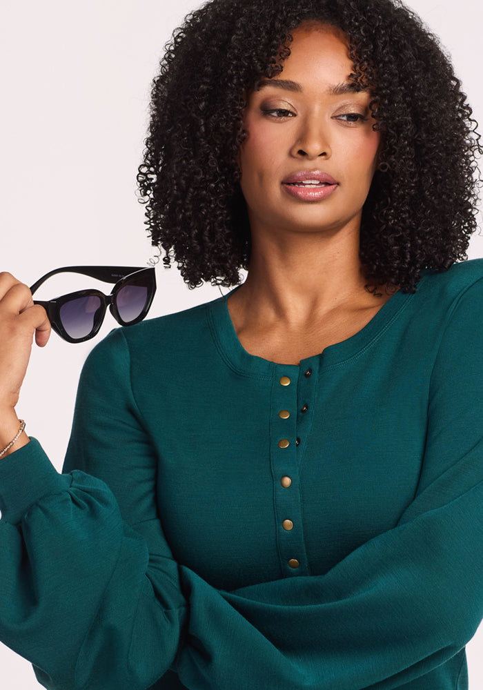 A woman with curly hair wears the Woolx Charlotte Puff Sleeve Top in Riverbank Green, holding black sunglasses in her right hand and looking to the side with a neutral expression.