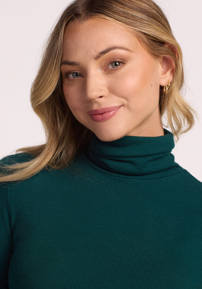A woman with wavy blonde hair, wearing the Woolx Sage Ribbed Turtleneck in Riverbank Green and gold hoop earrings, smiles softly at the camera against a light background.