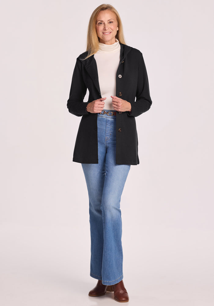 A woman with long blonde hair wears the Woolx Edna Hooded Cardigan in black, paired with a white turtleneck, blue jeans, and brown shoes, smiling against a plain light background.