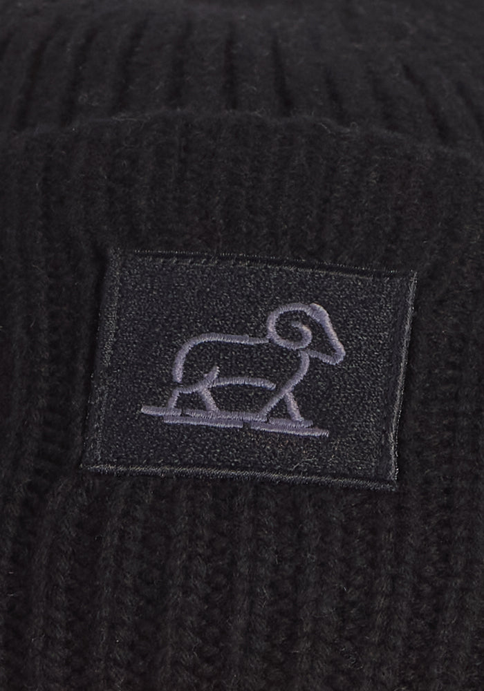 The Woolx Ria Chunky Beanie in black features a square patch with a light embroidered ram outline—a stylish and cozy winter accessory.