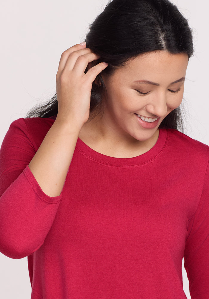 A woman with long dark hair smiles and looks down, touching her hair while wearing the Woolx Rissa A Line Dress in Poppy Spark. The plain light background highlights this stylish Merino wool piece, perfect for fall-winter layering.