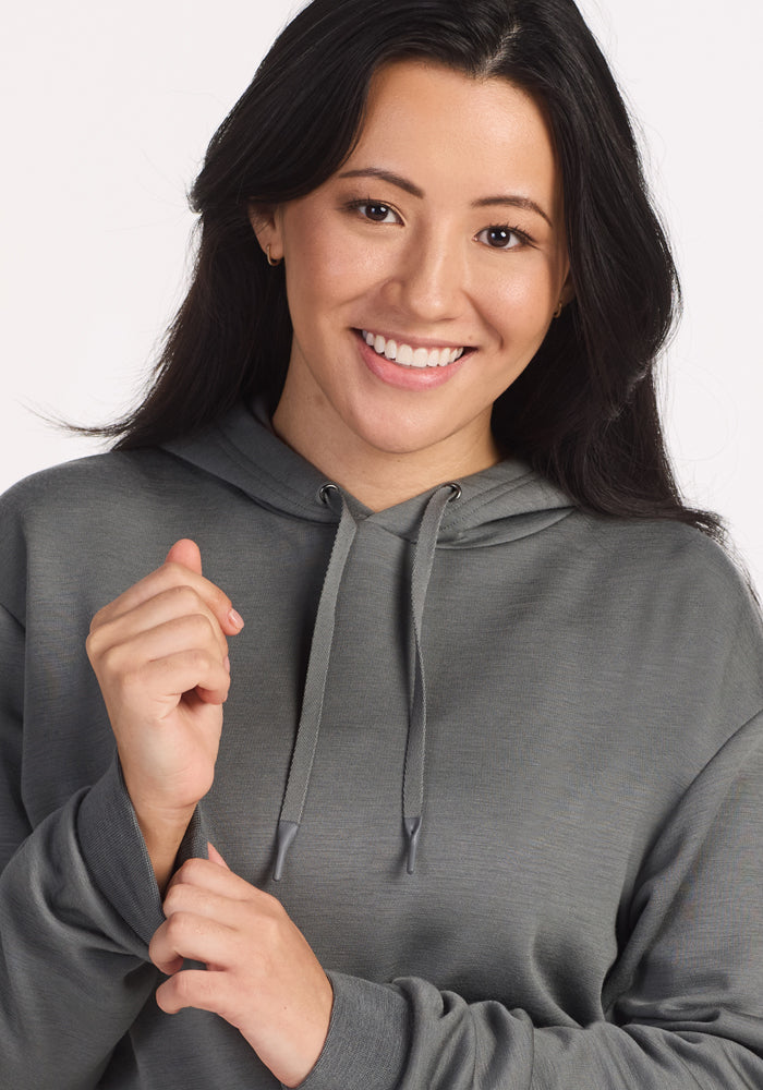 A woman with long dark hair smiles at the camera while wearing the Woolx Avery Hoodie in Sedona Sage, holding one sleeve with her left hand and standing against a plain white background.