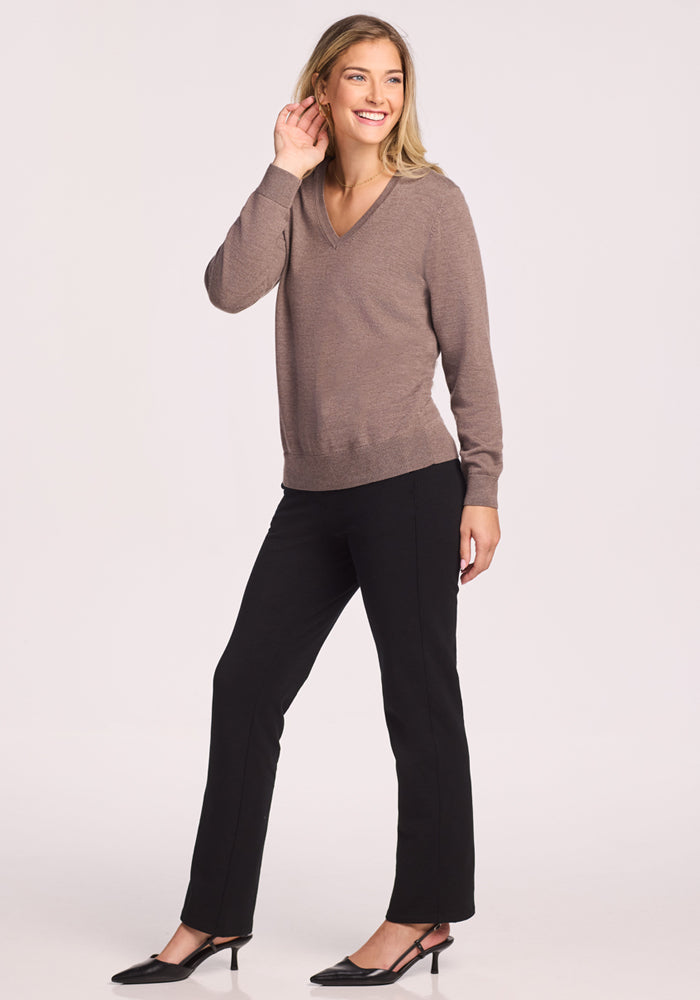 A woman with blond hair smiles, posing against a plain light background in the Woolx Francesca Sweater in Simply Taupe, paired with black pants and black heels. One hand is near her ear as she stands confidently.